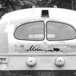 May include: Black and white image of the back of a vintage bus. The word "Moin" is written in cursive, with a paper boat sailing on a wavy line below it. The bus has two windows and a weathered, distressed appearance.