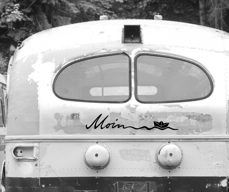 May include: Black and white image of the back of a vintage bus. The word "Moin" is written in cursive, with a paper boat sailing on a wavy line below it. The bus has two windows and a weathered, distressed appearance.