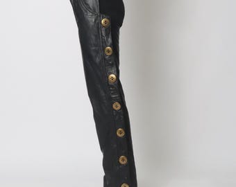 May include: A black short-sleeved top paired with black leather trousers featuring a side panel adorned with a vertical row of large, circular gold-toned buttons running from the waist down to the hem of the trouser leg.