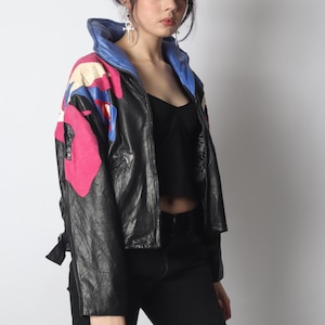 May include: A black leather jacket featuring a vibrant multi-colored floral pattern on the shoulders and sleeves. The jacket has a high, folded collar lined with blue leather, a front zipper closure, and long sleeves with a structured, boxy fit.