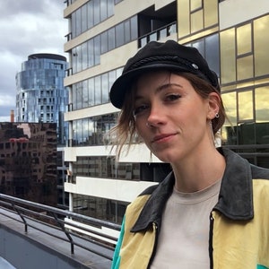 May include: A person wearing a black newsboy cap with a braided band, a yellow and teal jacket, and a white t-shirt. The person is standing in front of a building with large windows.