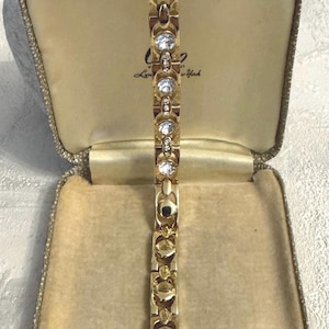 May include: A gold bracelet with clear gemstones in a repeating design, presented in an open, vintage jewellery box. The box has a cream interior and gold trim, with the text "London by New York". The bracelet is a classic piece.