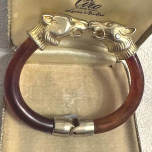 Puede incluir: Un brazalete vintage con una banda circular marrón y un cierre plateado. El brazalete presenta cabezas de león doradas en cada extremo, enfrentadas. Se presenta en una caja color crema con el texto "Ciro of Bond Street London & New York".