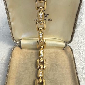 Puede incluir: Una pulsera de eslabones dorados con segmentos metálicos lisos y pulidos que se alternan con eslabones rectangulares incrustados con brillantes transparentes. La pulsera se presenta en un joyero texturizado con forro beige y el texto New York.