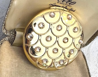 May include: A round gold-tone compact mirror featuring a scalloped scale pattern with embedded clear crystals. The compact is displayed inside a beige textured case with the text Ciro of Bond Street London & New York printed on the interior lining.