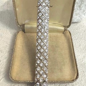 May include: A silver bracelet with a geometric pattern, set with many clear, sparkling rhinestones. The bracelet is presented in a vintage, gold-toned jewellery box with a soft, velvet interior.