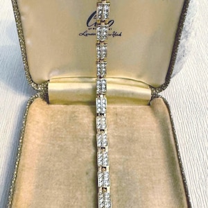 May include: A gold bracelet with rectangular links, each set with clear crystals. The bracelet is presented in an open, vintage jewellery box with a cream interior and gold trim. The box has the text "London New York".