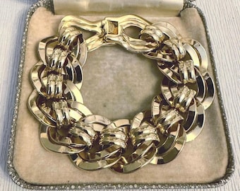 Puede incluir: Un brazalete dorado con eslabones grandes e interconectados, presentado en un joyero vintage. El brazalete presenta un cierre seguro y un acabado pulido, que refleja la luz. El joyero tiene un interior beige suave y un borde decorativo con cuentas.