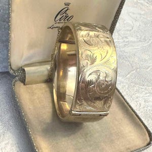May include: A gold-toned hinged bangle bracelet featuring an intricate engraved scrollwork pattern across its surface. The bracelet includes a thin safety chain and is presented inside a textured, cream-lined jewelry box with the Ciro of Bond Street logo.