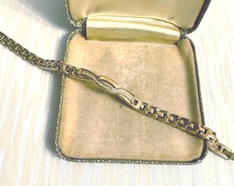 Puede incluir: Una pulsera dorada con un diseño de eslabones y una sección central adornada con pequeñas piedras preciosas transparentes. La pulsera se encuentra dentro de un joyero vintage con interior crema y borde decorado con gemas.