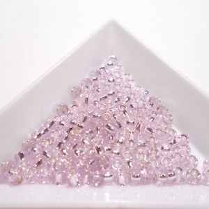 Puede incluir: Un montón de pequeñas cuentas de semillas de vidrio de color rosa claro con acentos plateados. Las cuentas están dispuestas en forma triangular sobre una superficie blanca.