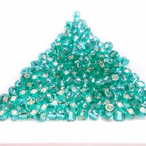 Puede incluir: Un montón de cuentas de semillas de vidrio verde turquesa con acentos plateados. Las cuentas están dispuestas en forma triangular.