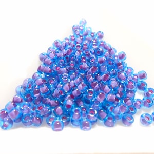 Puede incluir: Un montón de pequeñas cuentas de vidrio azul claro y morado. Las cuentas son translúcidas y tienen un ligero brillo iridiscente.