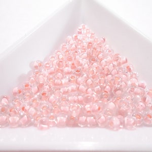 Può includere: Un mucchio di piccole perline di seme rosa traslucide. Le perline sono disposte in un mucchio sciolto su una superficie bianca.