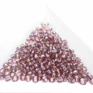 Puede incluir: Un montón de pequeñas cuentas de semillas de vidrio rosa iridiscente. Las cuentas son redondas y tienen un brillo metálico.