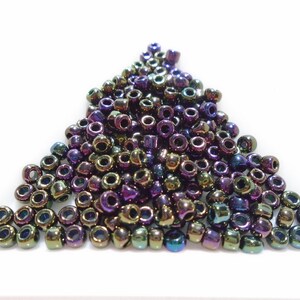 Puede incluir: Un montón de cuentas de vidrio iridiscente en tonos de morado, verde y dorado. Las cuentas son pequeñas y redondas, y están dispuestas en una forma triangular suelta.