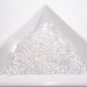 Puede incluir: Un montón de cuentas de semillas de vidrio transparente en un plato triangular blanco. Las cuentas son pequeñas y redondas, y son perfectas para hacer joyas.