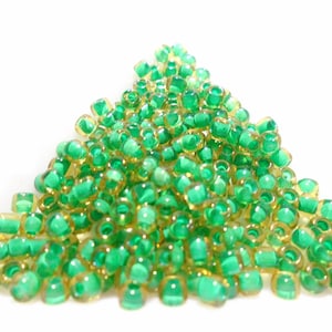 Puede incluir: Un montón de cuentas de semillas verdes y doradas. Las cuentas son pequeñas y redondas, y están dispuestas en un montón suelto.