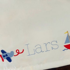 May include: White embroidered baby blanket with the name "Lars" in blue thread. The blanket features a blue airplane with a red tail and a red sailboat with a blue sail.