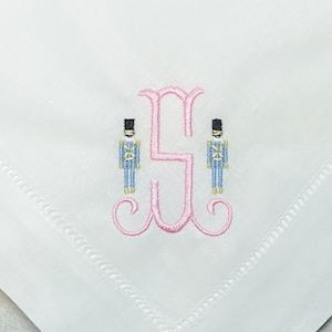 May include: White handkerchief featuring a pink embroidered initial flanked by two toy soldier figures. The soldiers wear blue and gold uniforms with black hats. The initial is in a decorative script font.