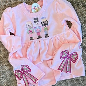 May include: Pink sweatshirt dress with a nutcracker design and pink and green plaid bows on the sleeves. The dress has a round neckline and long sleeves. The text "Bubbly & Red" is on the tag.