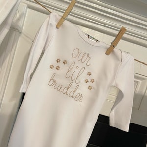 Op de afbeelding: Witte baby romper met lange mouwen met gouden borduurwerk met de tekst "our lil brudder" met poot afdrukken rond de tekst.
