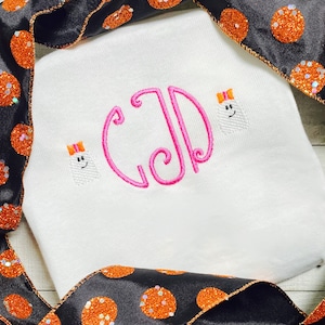 Puede incluir: Camiseta blanca con un monograma rosa "CJD" y dos fantasmas bordados con lazos rosas. La camiseta está sobre una cinta negra y naranja con purpurina.