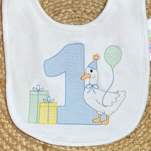 May include: A white bib with blue and green embroidery. The bib features a goose holding a balloon, a number one, and two presents. The text "One" is embroidered on the bib.