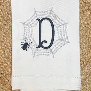 Puede incluir: Toalla decorativa blanca con un diseño de telaraña gris, una araña negra y la letra "D" bordada en azul oscuro. La toalla tiene un patrón texturizado y un borde decorativo.