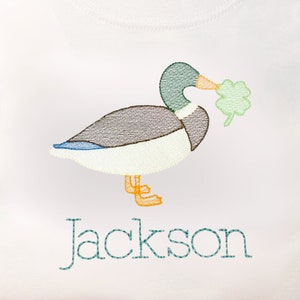May include: A white t-shirt with an embroidered image of a green duck holding a shamrock in its beak. The name "Jackson" is embroidered below the duck.