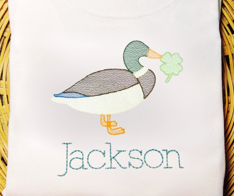 May include: A white t-shirt with an embroidered image of a green duck holding a shamrock in its beak. The name "Jackson" is embroidered below the duck.