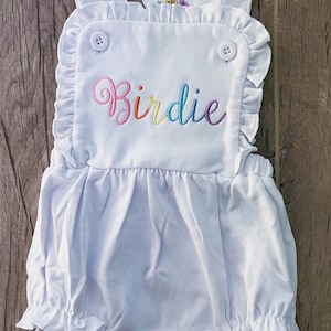 May include: White baby romper with a rainbow embroidered name "Birdie" on the front. The romper has ruffles around the neckline and the bottom.