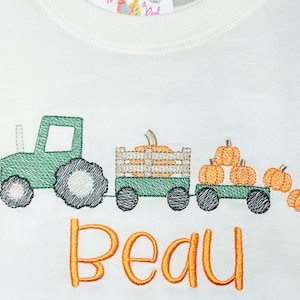 Puede incluir: Una camiseta blanca con un diseño bordado de un tractor verde tirando de un vagón lleno de calabazas naranjas. El nombre "Beau" está bordado en naranja debajo del tractor.