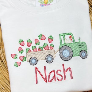 Op de afbeelding: Een wit T-shirt met geborduurde rode aardbeien, een groene tractor die een wagen vol aardbeien trekt, en de naam "Nash" in rode borduurwerk.