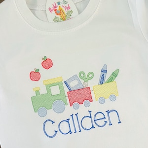 Puede incluir: Una camiseta blanca con un diseño bordado de un tren que lleva útiles escolares. El tren es verde, rojo y amarillo. El tren lleva un libro, tijeras, crayones y un lápiz. El nombre "Callden" está bordado debajo del tren.