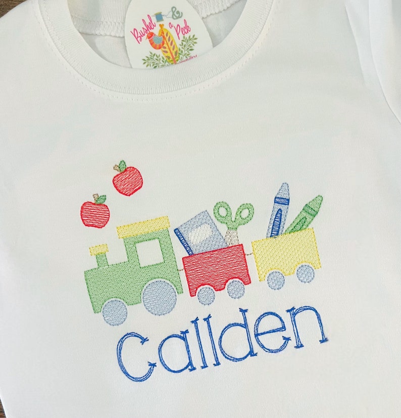 May include: A white t-shirt with an embroidered design of a train carrying school supplies. The train is green, red, and yellow. The train is carrying a book, scissors, crayons, and a pencil. The name "Callden" is embroidered below the train.