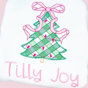 Peut inclure: Un arbre de Noël brodé blanc avec un nœud rose et un motif écossais vert et blanc. L'arbre est décoré de lumières roses et le nom "Tilly Joy" est brodé en dessous.