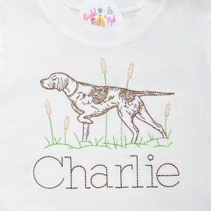 May include: White t-shirt with brown embroidered image of a dog with the name "Charlie" underneath.
