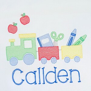 May include: Embroidered design of a green and yellow toy train carrying a red box, a blue book, a pair of scissors, a blue crayon, and a green crayon. Two red apples are above the train. The name "Callen" is embroidered in blue below the train.
