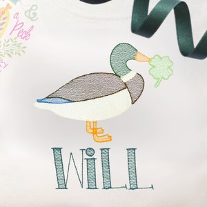 May include: Embroidered design of a green duck holding a shamrock in its beak, with the name "WILL" embroidered below.