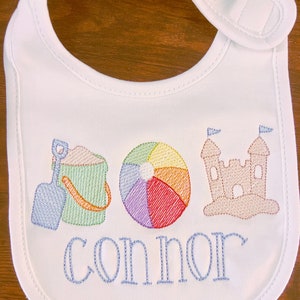May include: White bib with blue embroidery of a beach pail, shovel, beach ball, sandcastle, and the name "Connor".