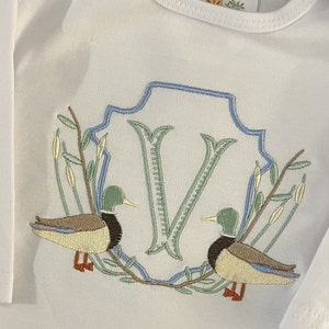 May include: Embroidered white baby onesie with a blue and green monogrammed letter V surrounded by two ducks and leafy branches.