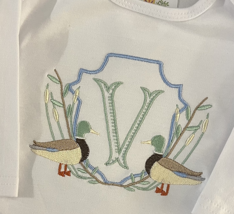 May include: Embroidered white baby onesie with a blue and green monogrammed letter V surrounded by two ducks and leafy branches.