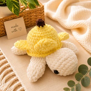 May include: A crocheted turtle toy in yellow and cream. The turtle has a yellow shell with a brown top, cream flippers, and a cream head with a black eye. A small card in the background reads "made with ♡".