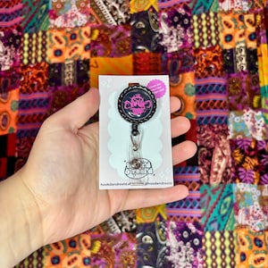 May include: A retractable badge reel featuring a black bottle cap top with a neon pink graphic of a stylized monkey face. The reel is attached to a white display card with the text hookdandreeld.etsy.com and hookdandreeld printed on it.
