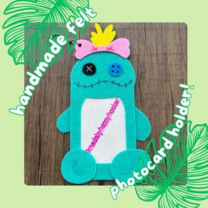 May include: Handmade felt photocard holder in the shape of a cartoon character. The teal-colored holder has a white center, a pink bow, and a yellow crown. The face has a stitched mouth, a black X, and a blue button eye. The text "handmade felt" and "Photocard holder!" are visible.