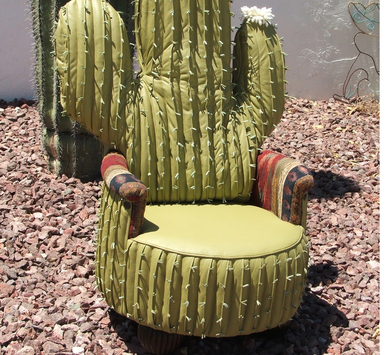 K&ouml;nnte beinhalten: Ein gr&uuml;ner Kaktus-f&ouml;rmiger Stuhl mit einer wei&szlig;en Bl&uuml;te oben. Der Stuhl hat einen braunen und roten gemusterten Stoff an den Armlehnen und ein gr&uuml;nes Kissen.