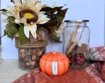 May include: A pumpkin-shaped candle with a lid, in a bright orange color, with the text "Pumpkin Spice" on the front. The candle is sitting on a brown and white blanket, with a sunflower and a glass jar in the background.