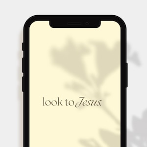 May include: A black smartphone displays a light yellow screen with the text "look to Jesus" in a brown script font. A floral shadow is visible on the screen. The phone is set against a white background.