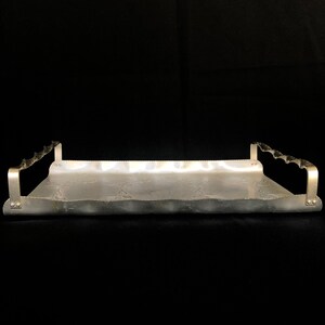 May include: A silver serving tray with decorative embossed details and two handles on each side. The tray is made of aluminum and has a slightly wavy edge.